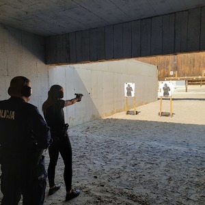 Policjantka strzela z broni krótkiej do tarczy-na nią stoi instruktor strzelectwa