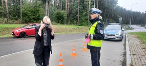 Kobieta w alkogoglach porusza się pomiędzy pachołkami pod opieka policjantki