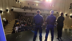 Na zdjęciu umundurowani policjanci, dyrektor MDK oraz w tle uczniowie na widowni