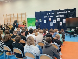 Na zdjęciu widać przedstawicieli innych zawodów, uczniów oraz policjanta prowadzącego pogadankę